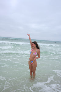 Woman in a colorful bikini standing in shallow ocean water with arms raised on a cloudy day.