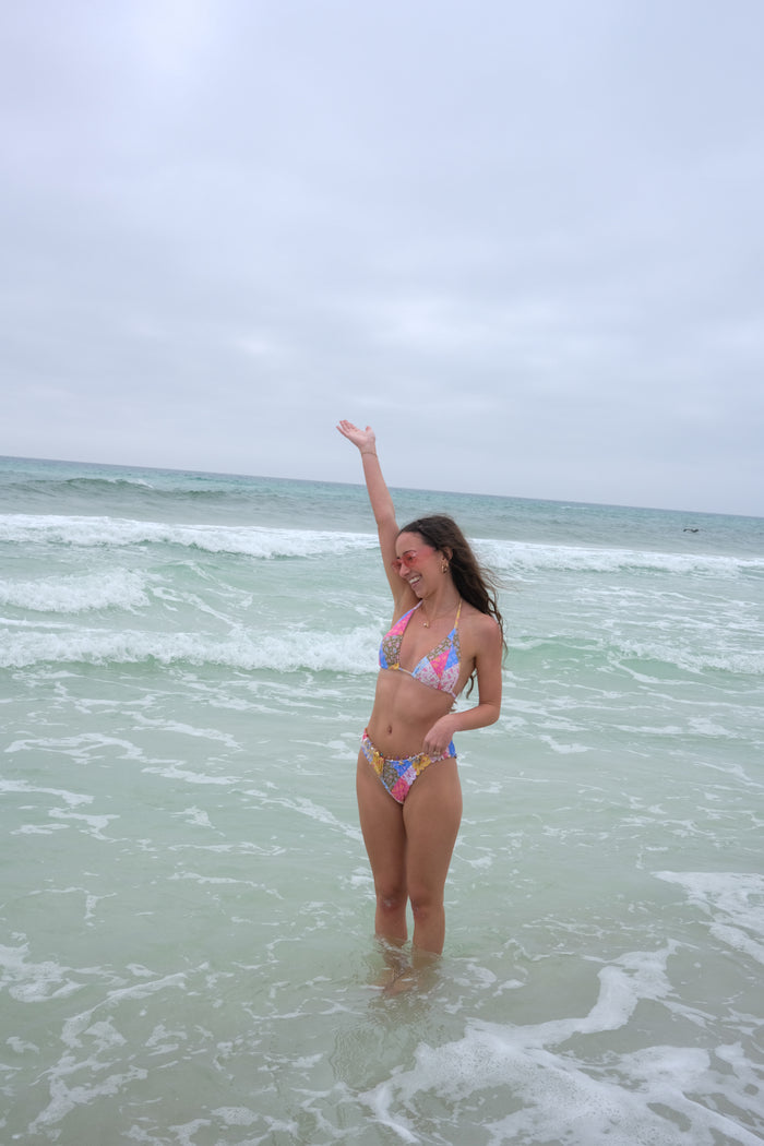 Woman in a colorful bikini standing in shallow ocean water with arms raised on a cloudy day.