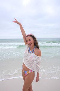 Woman in a colorful bikini and white cover-up standing on a beach with ocean waves in the background.