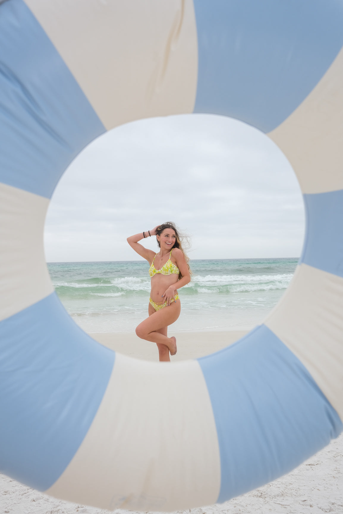 Woman in a yellow bikini standing on a beach with a blue and white striped float in the foreground.