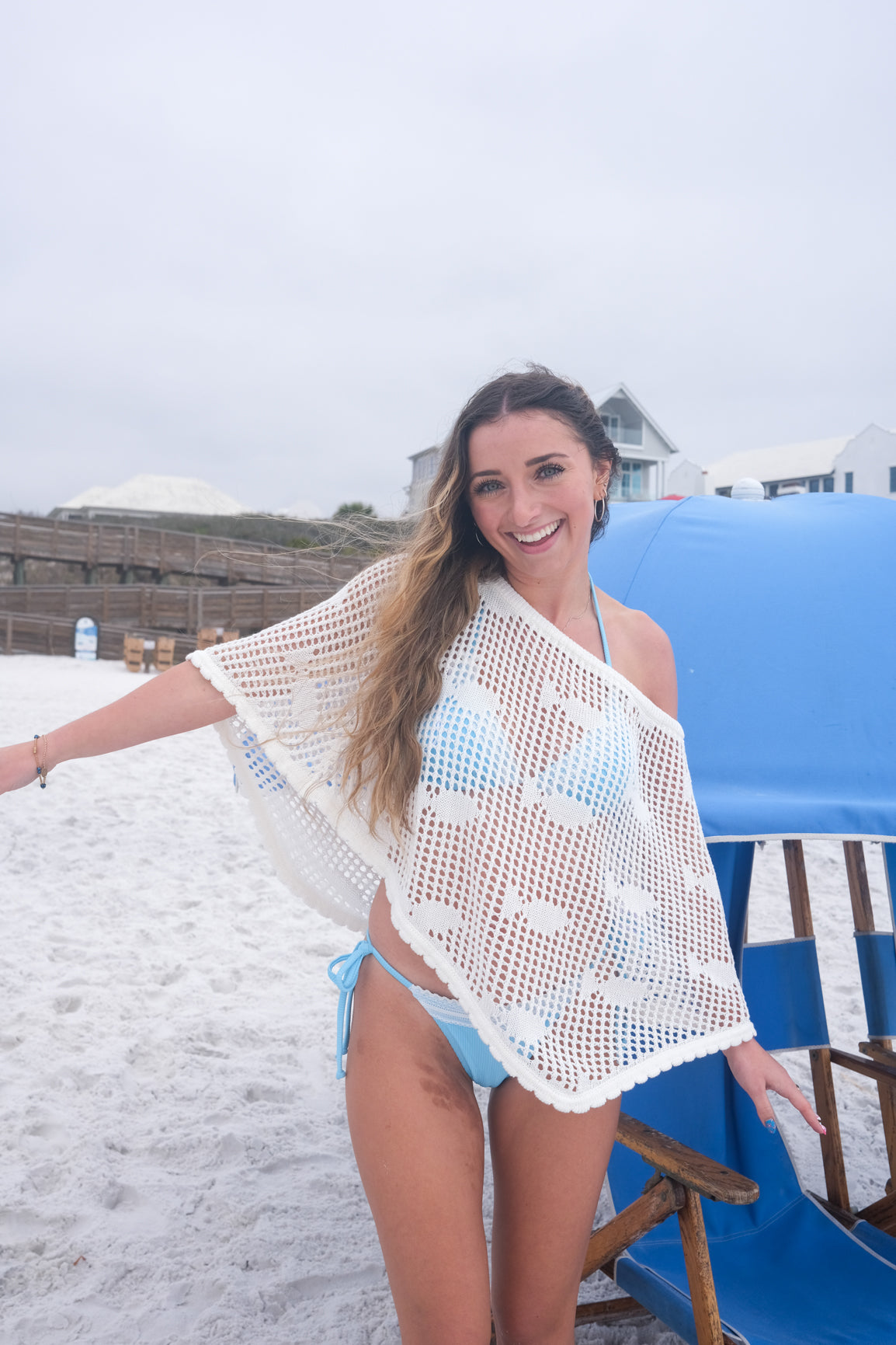 Woman on a beach wearing a white crochet cover-up and blue bikini, sitting on a blue chair.