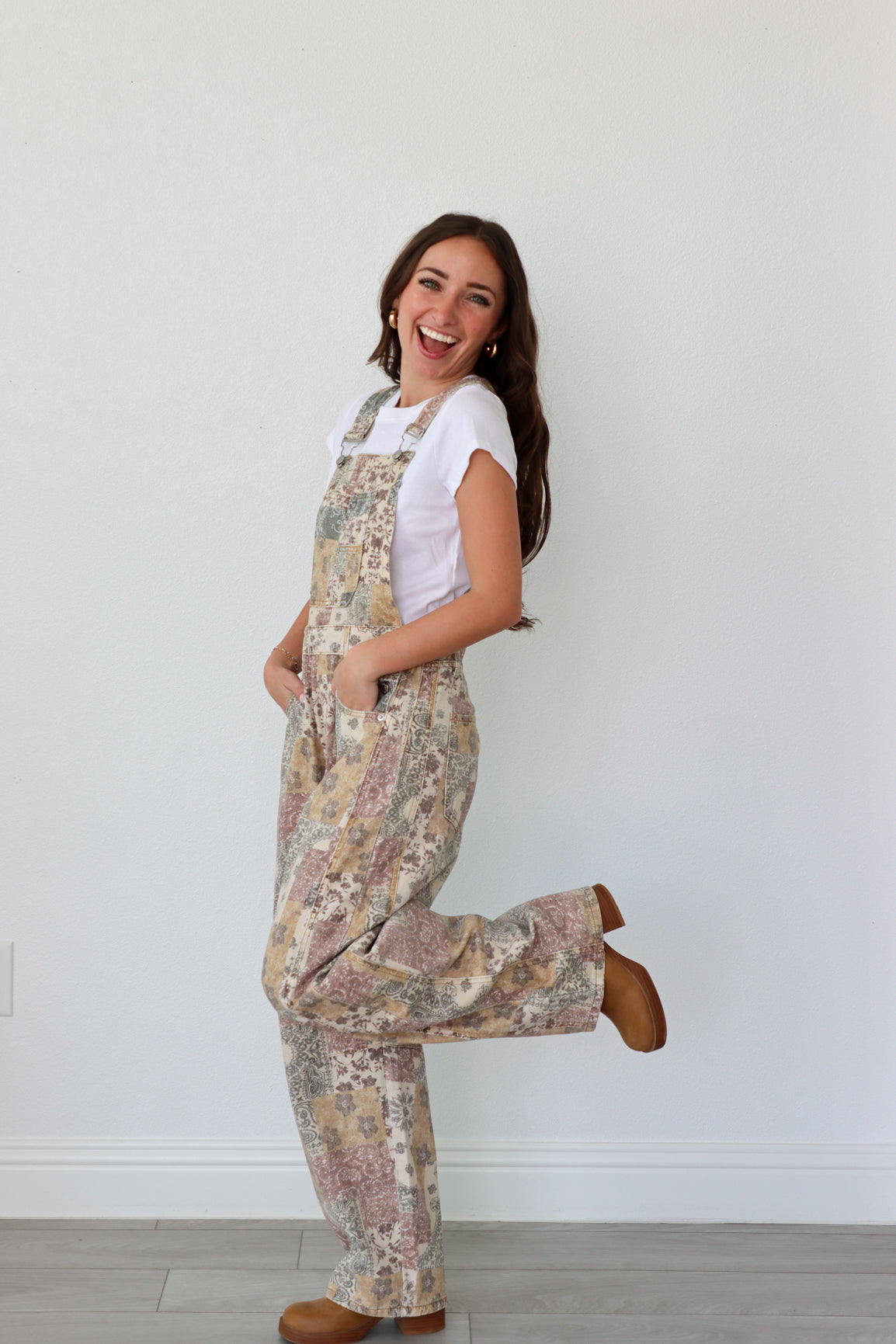 Woman wearing overalls and white shirt, posing against a white wall.