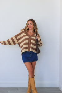 Woman wearing a brown and beige striped sweater with blue shorts and tan boots against a white wall.