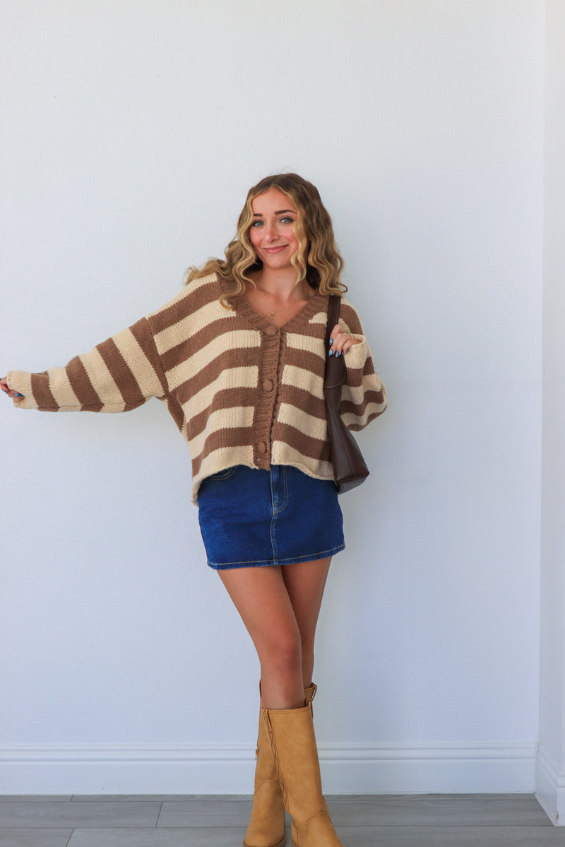 Woman wearing a brown and beige striped sweater with blue shorts and tan boots against a white wall.