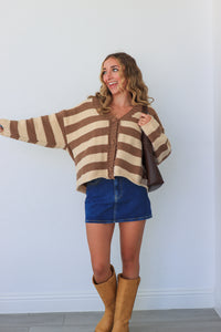 Woman wearing a brown and beige striped sweater, blue shorts, and tan boots against a white wall.