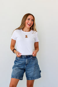 Woman wearing a white t-shirt with a logo and denim shorts against a plain background