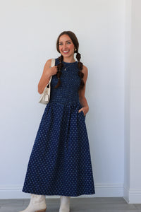 Woman wearing a blue polka dot dress with white boots against a white wall.