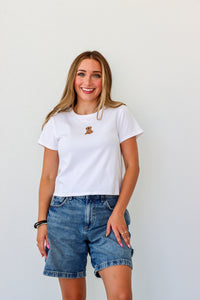 Woman wearing a white t-shirt with a logo and denim shorts against a plain background
