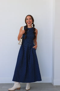 Woman wearing a blue polka dot dress with white boots against a white wall.