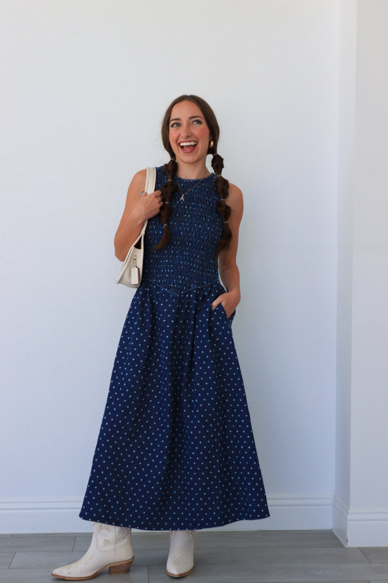 Woman wearing a blue polka dot dress with white boots against a white wall.