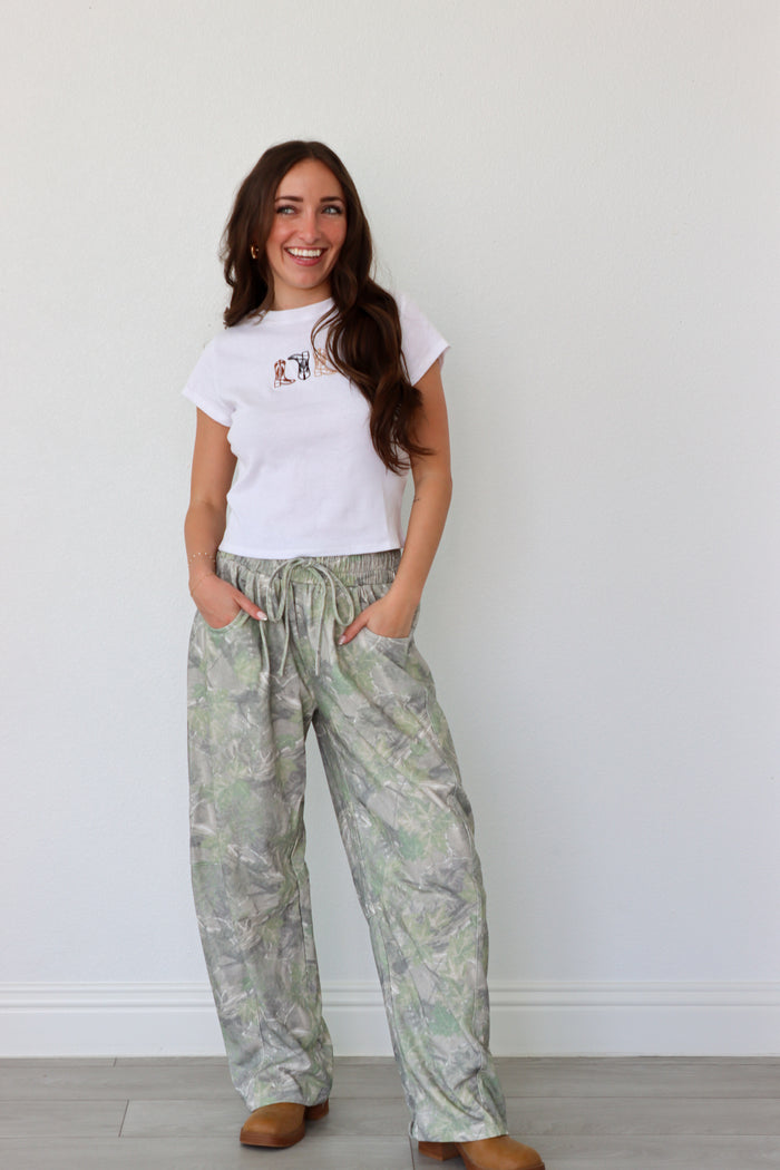 Woman wearing a white t-shirt with a graphic design and light-colored patterned pants against a plain background