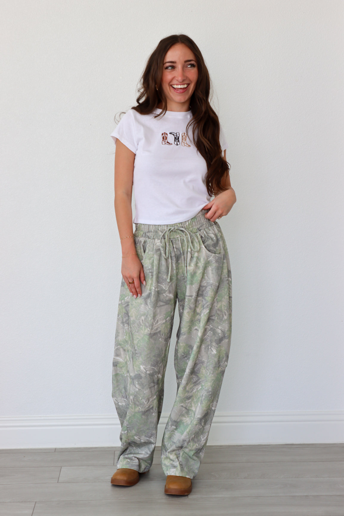 Woman wearing a white t-shirt with a graphic design and light green patterned pants against a white wall.