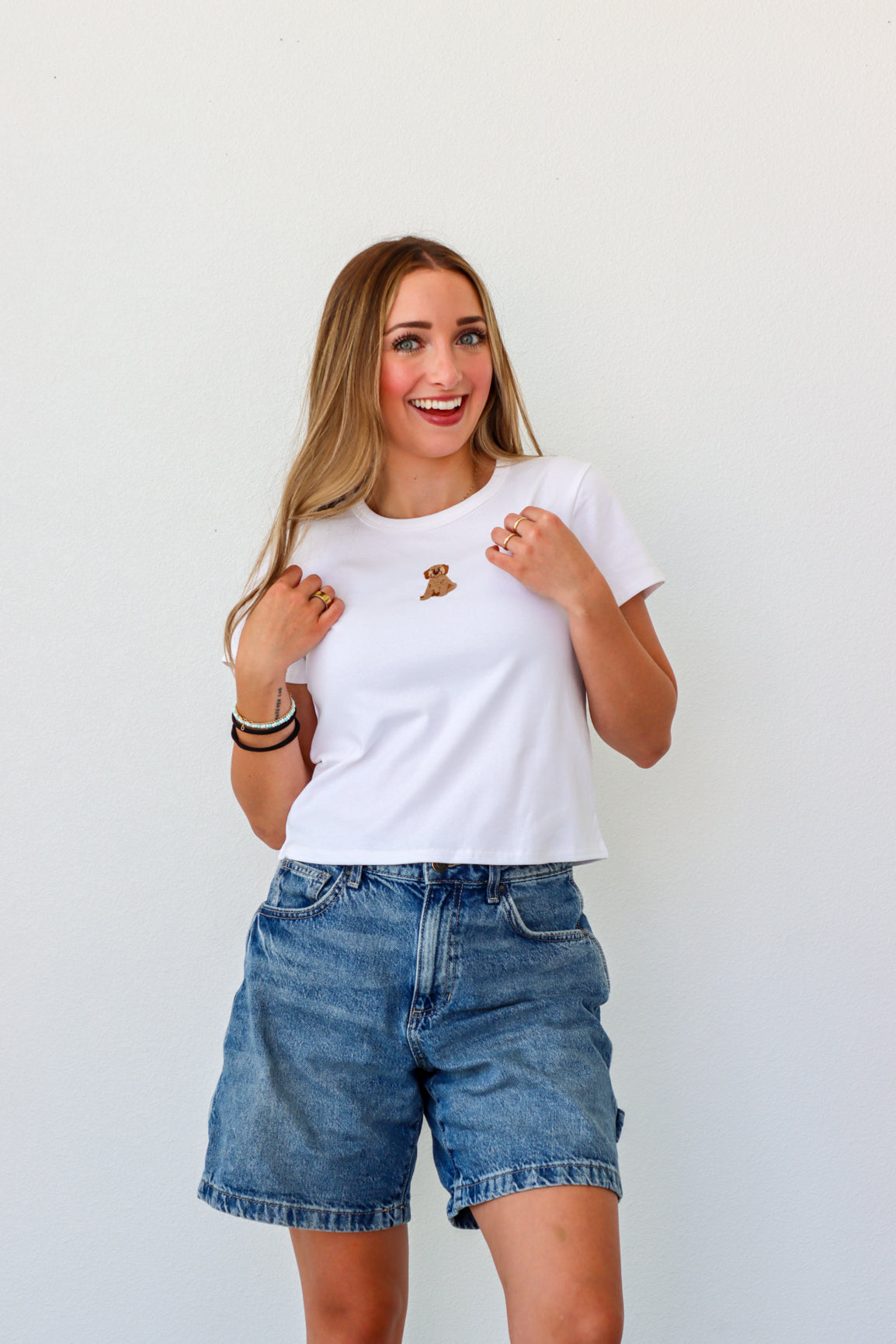 Woman wearing a white t-shirt with a logo and denim shorts against a plain background