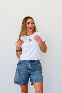 Woman wearing a white t-shirt with a logo and denim shorts against a plain background