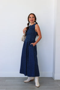 Woman wearing a blue polka dot dress with white boots against a white wall.
