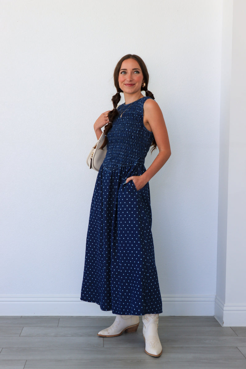 Woman wearing a blue polka dot dress with white boots against a white wall.