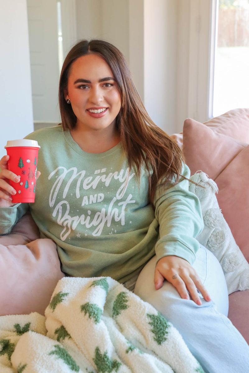 girl wearing light green crewneck sweatshirt with "be merry and bright" graphic