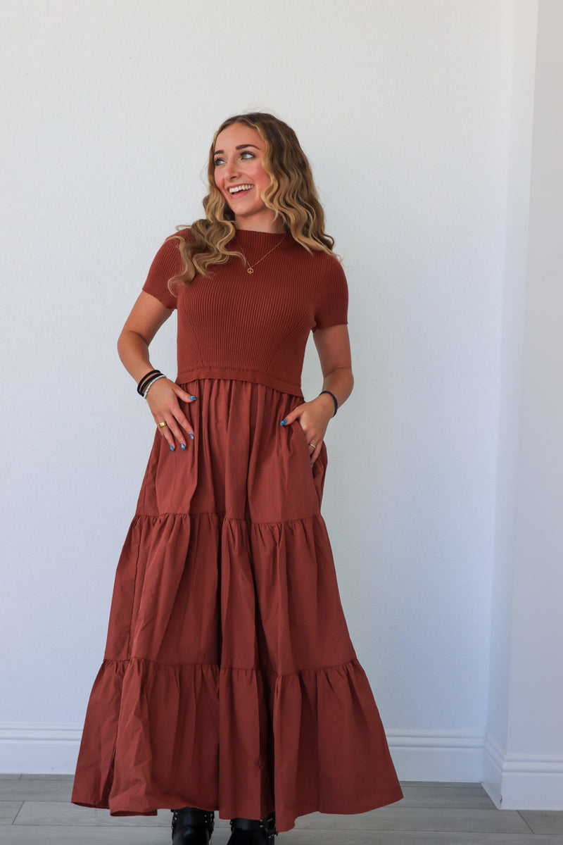 Woman wearing a long rust-colored dress against a white wall.
