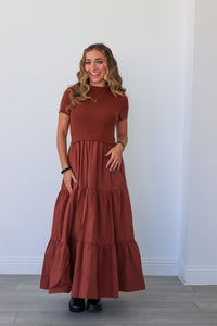 Woman wearing a long rust-colored dress against a white wall.