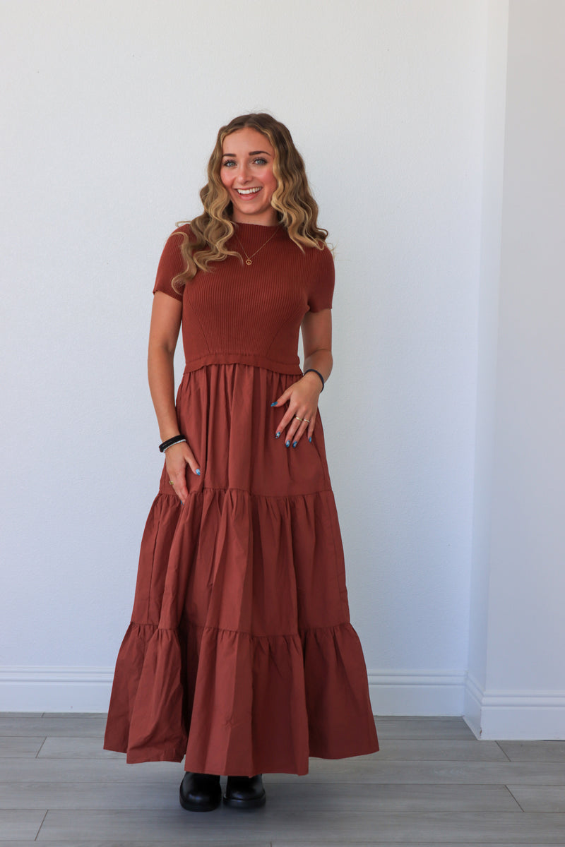 Woman wearing a long rust-colored dress against a white wall.
