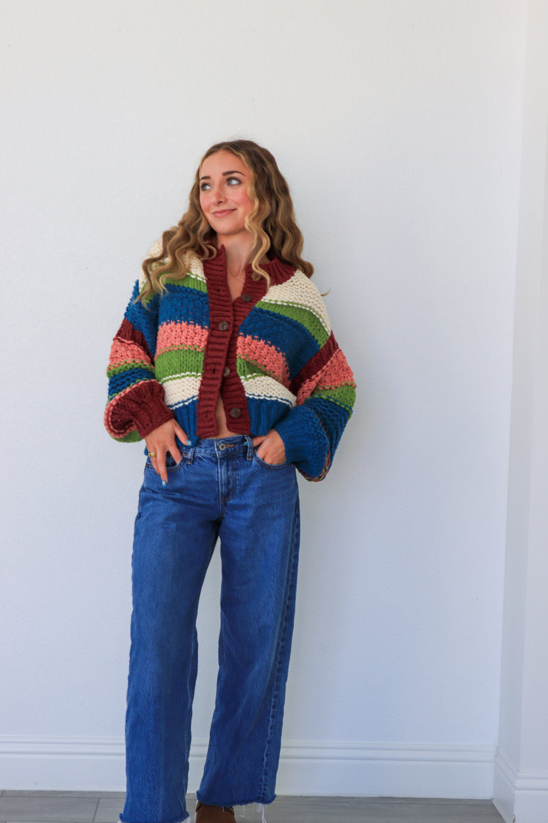 Woman wearing a colorful striped sweater and blue jeans against a white wall.