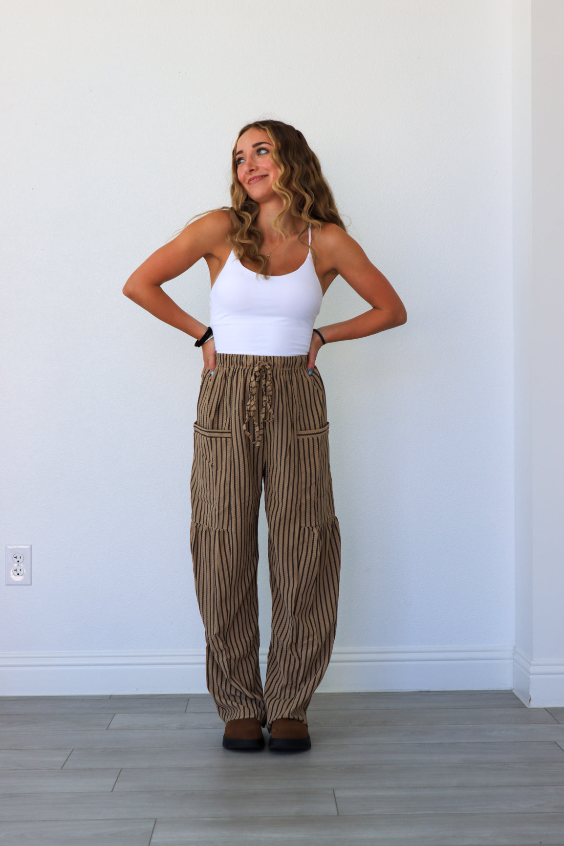 Woman wearing a white tank top and striped pants standing against a white wall.