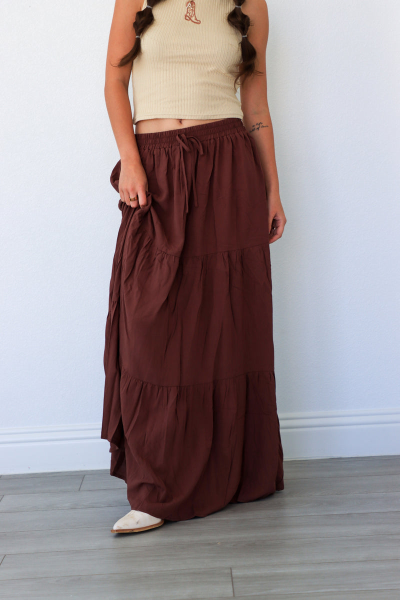 Person wearing a beige top and brown tiered skirt against a white wall.