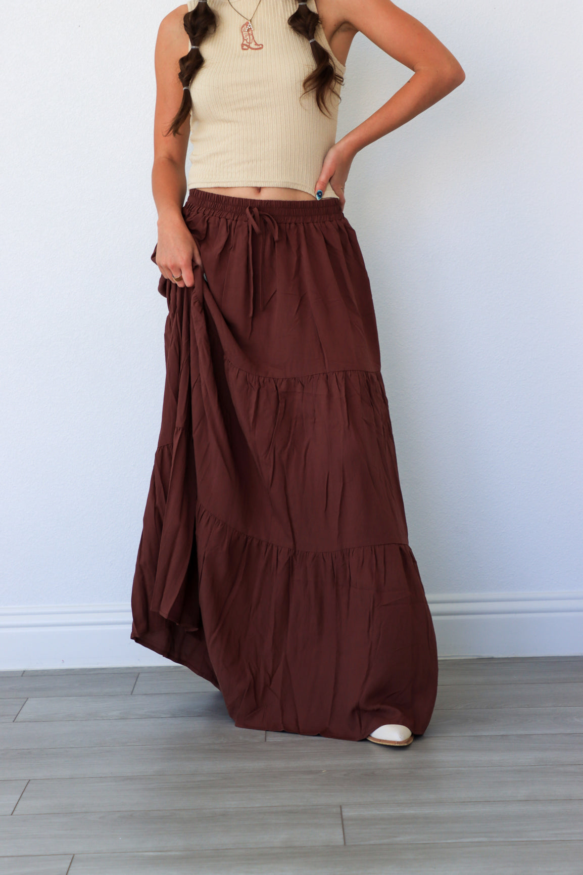 Woman wearing a long brown skirt and beige top against a white wall.
