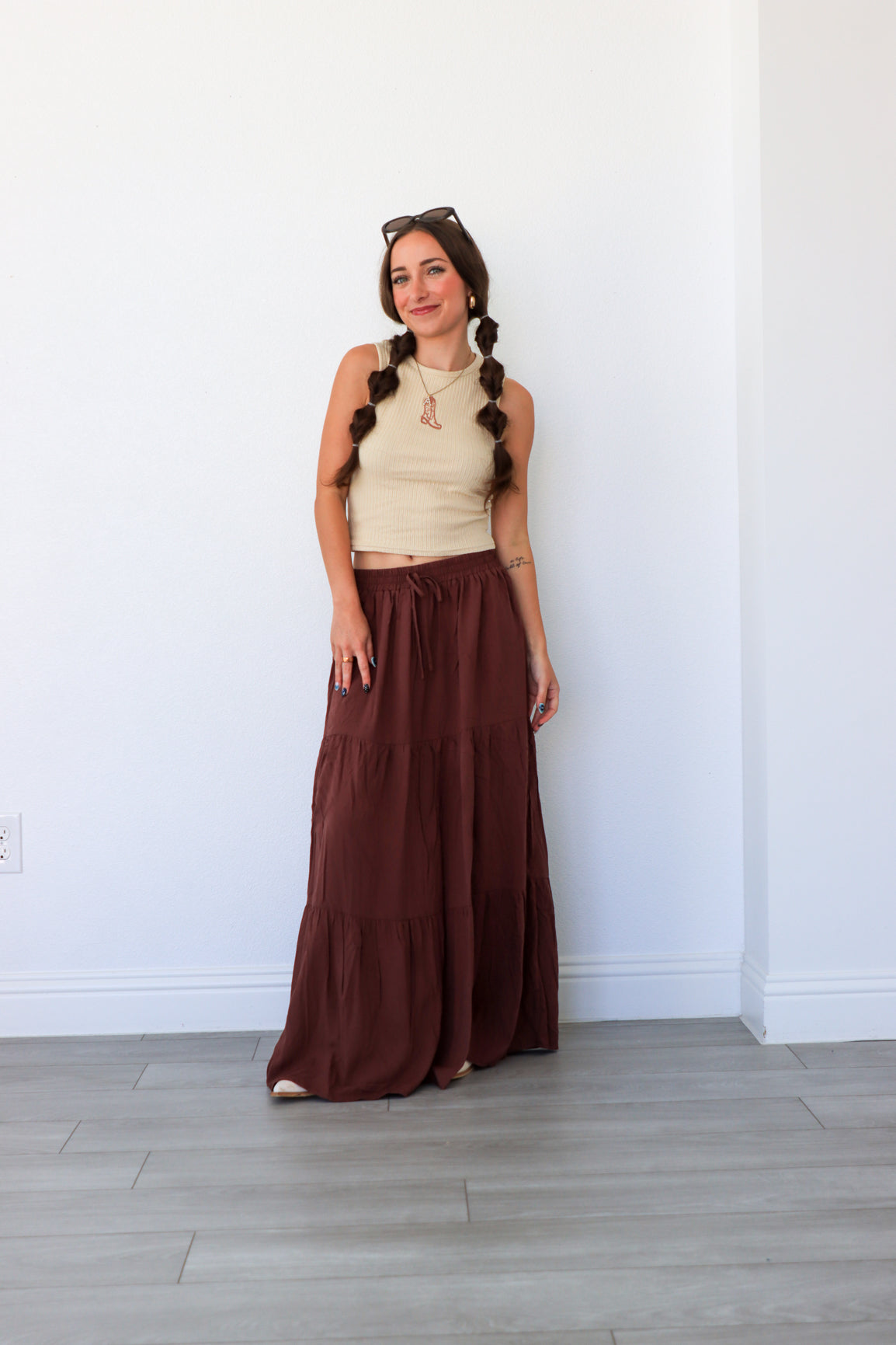 Woman wearing a beige top and brown long skirt standing against a white wall.