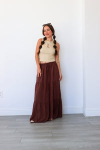 Woman wearing a beige top and brown long skirt standing against a white wall.