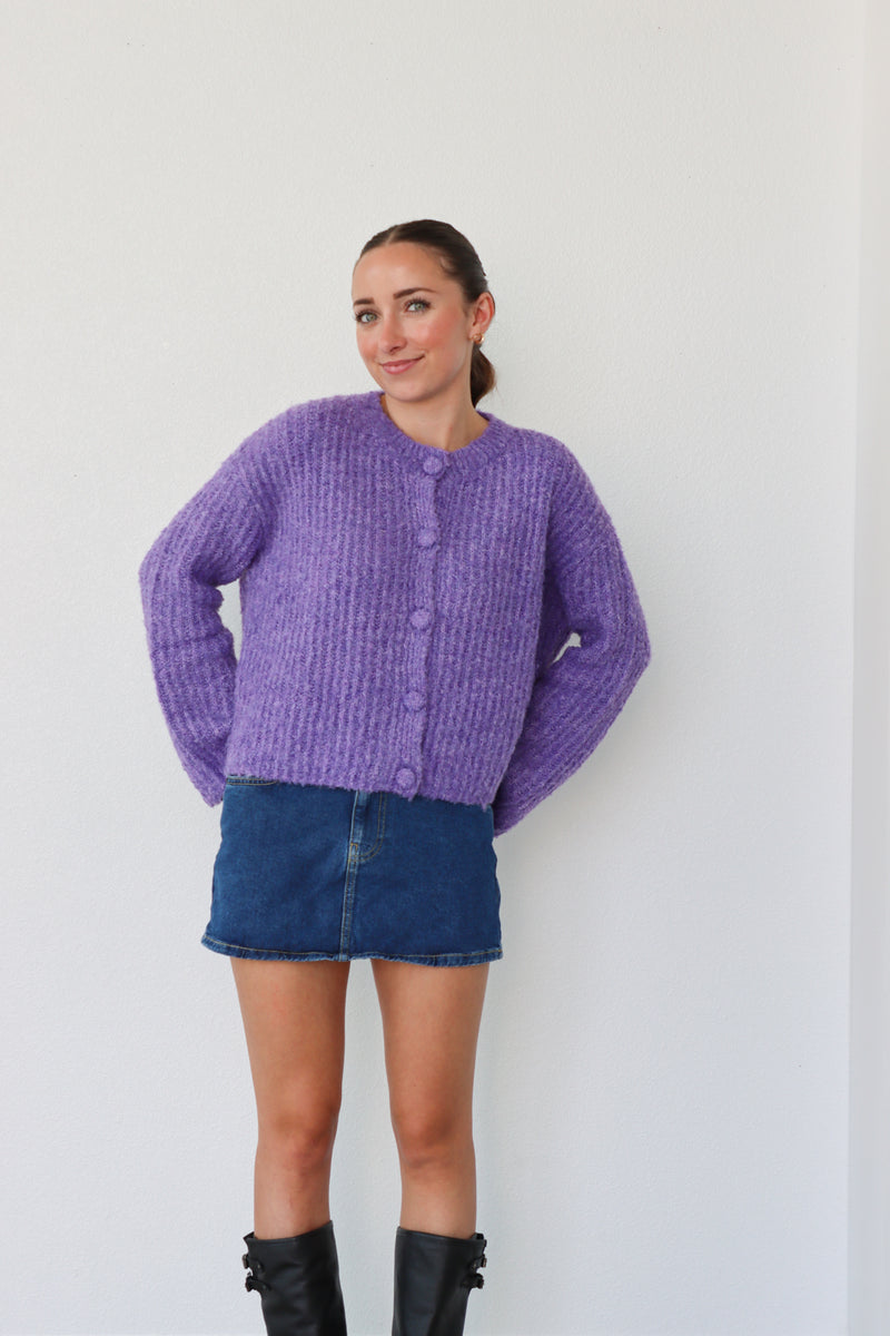 Woman wearing a purple knitted cardigan and denim skirt against a white background