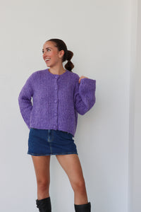 Woman wearing a purple cardigan, denim shorts, and black boots against a white background