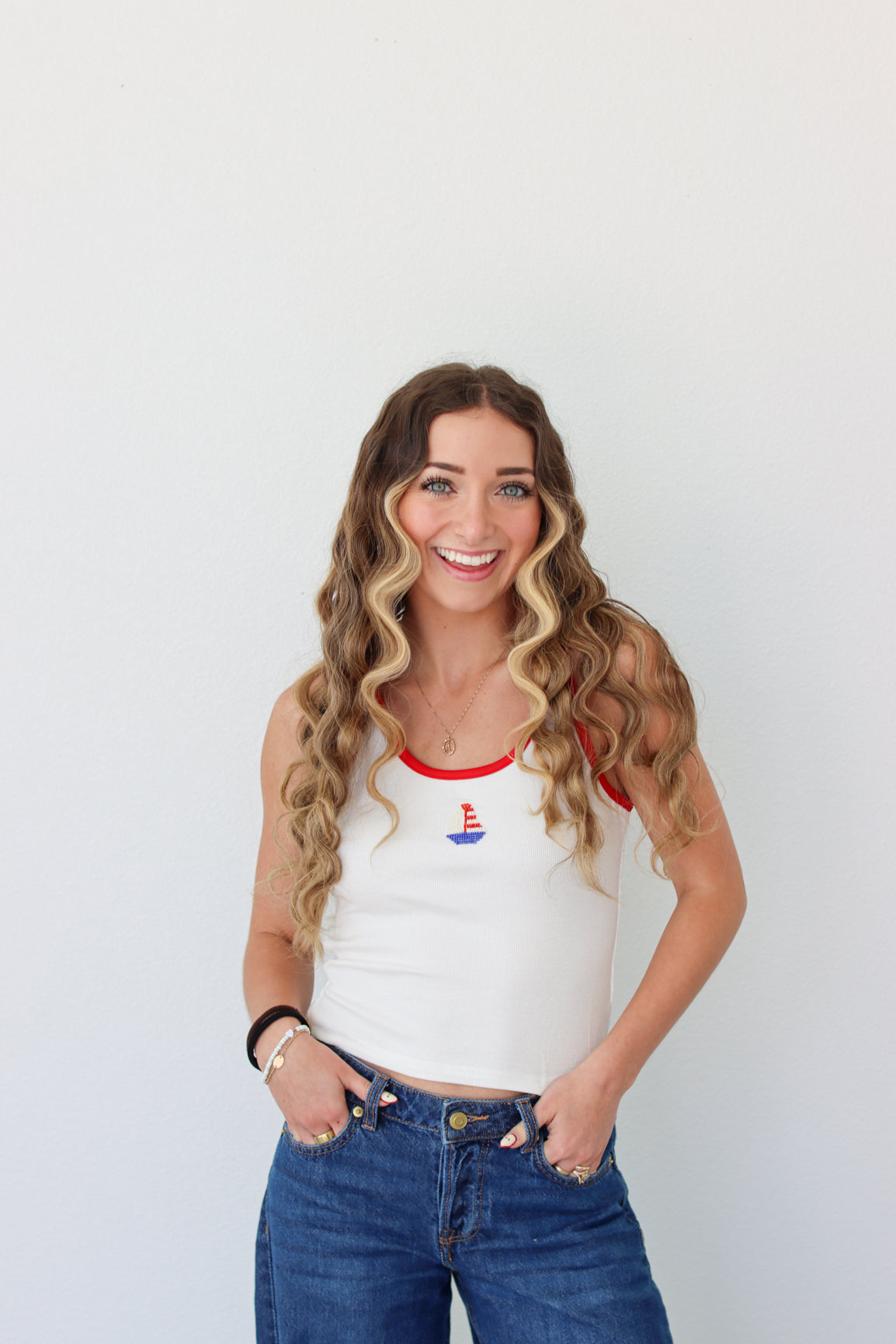 Woman wearing a white tank top with a red neckline and blue jeans against a plain background
