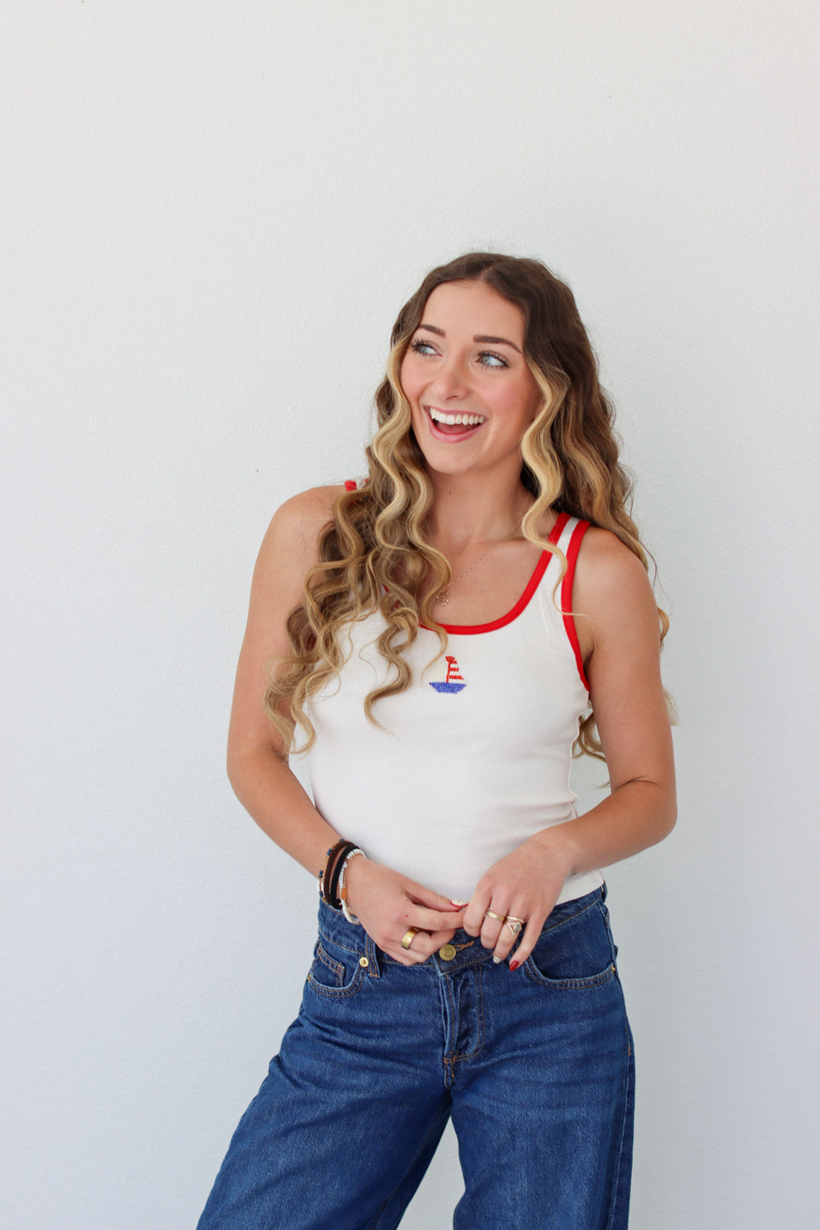 Woman wearing a white tank top with a red neckline and blue jeans against a plain background