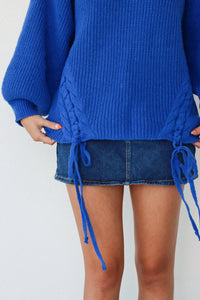 Person wearing a blue sweater with tie details and a denim skirt on a white background