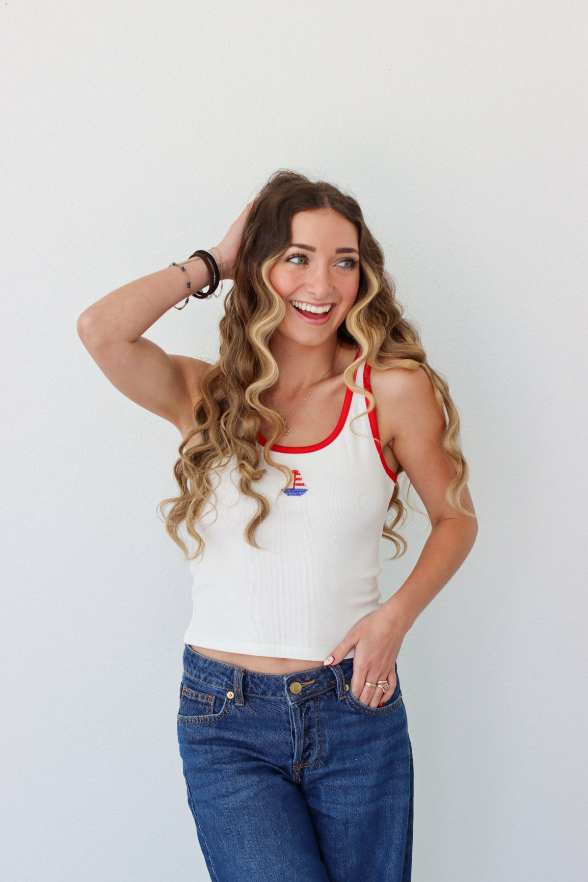 Woman wearing a white tank top with red trim and blue jeans against a plain background