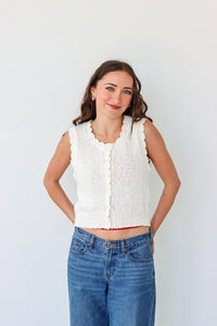 Woman wearing a white sleeveless knitted top and blue jeans against a white background