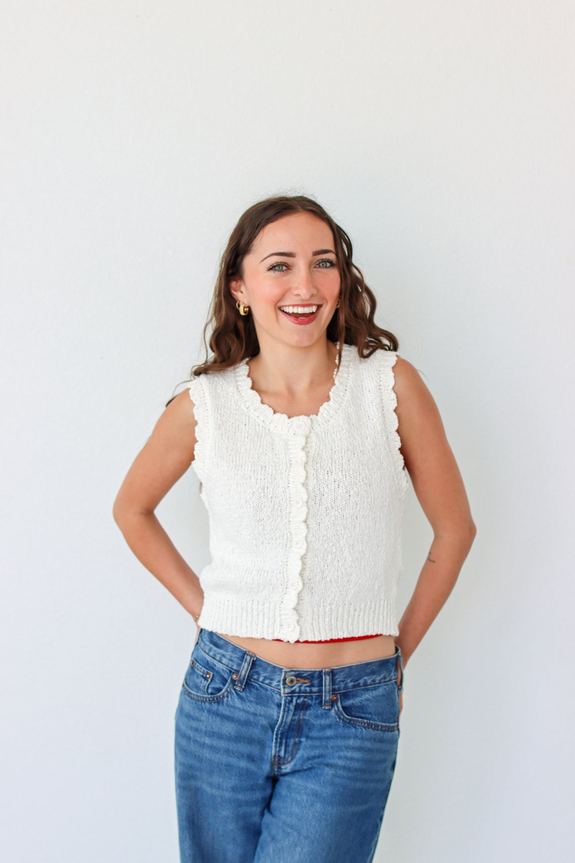Woman wearing a white sleeveless knitted top and blue jeans against a plain background
