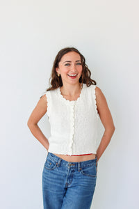 Woman wearing a white sleeveless knitted top and blue jeans against a plain background