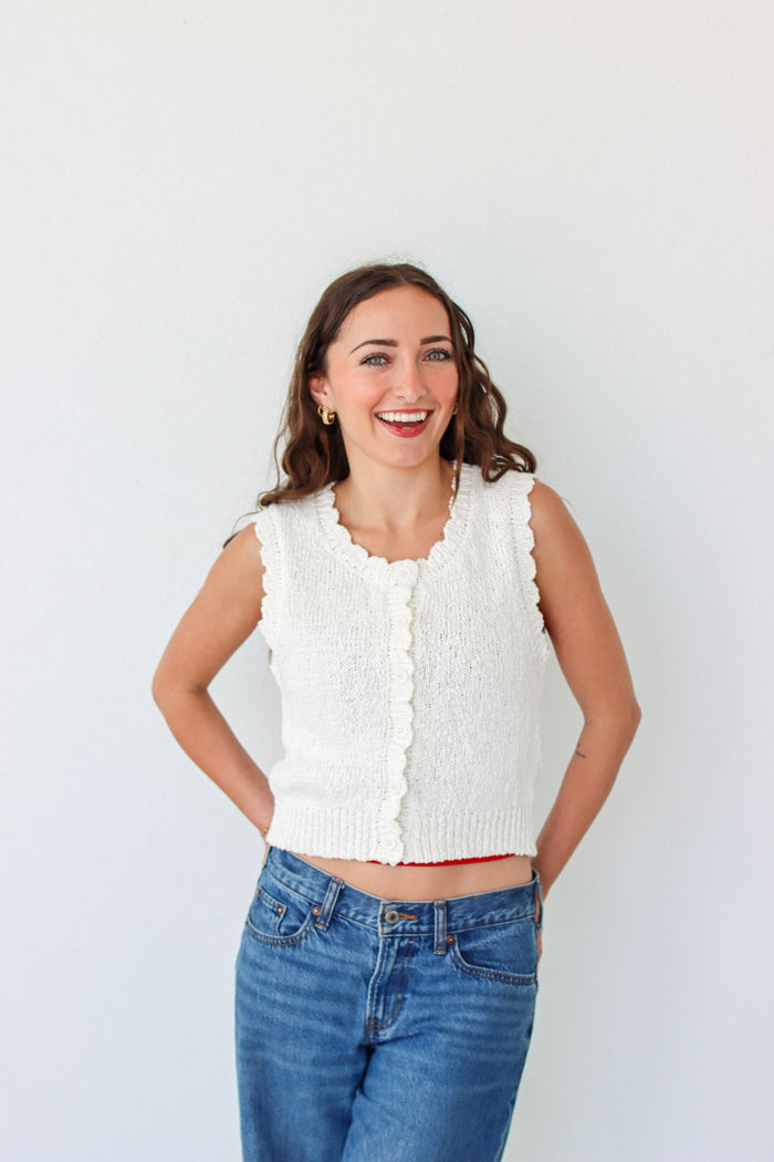 Woman wearing a white sleeveless knitted top and blue jeans against a plain background