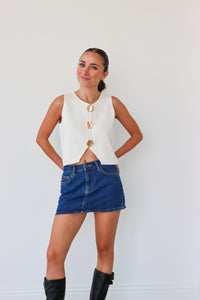 Woman wearing a white sleeveless top with gold buttons and a blue denim skirt against a white background