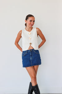 Woman wearing a white sleeveless top with button details and a blue denim skirt against a white background