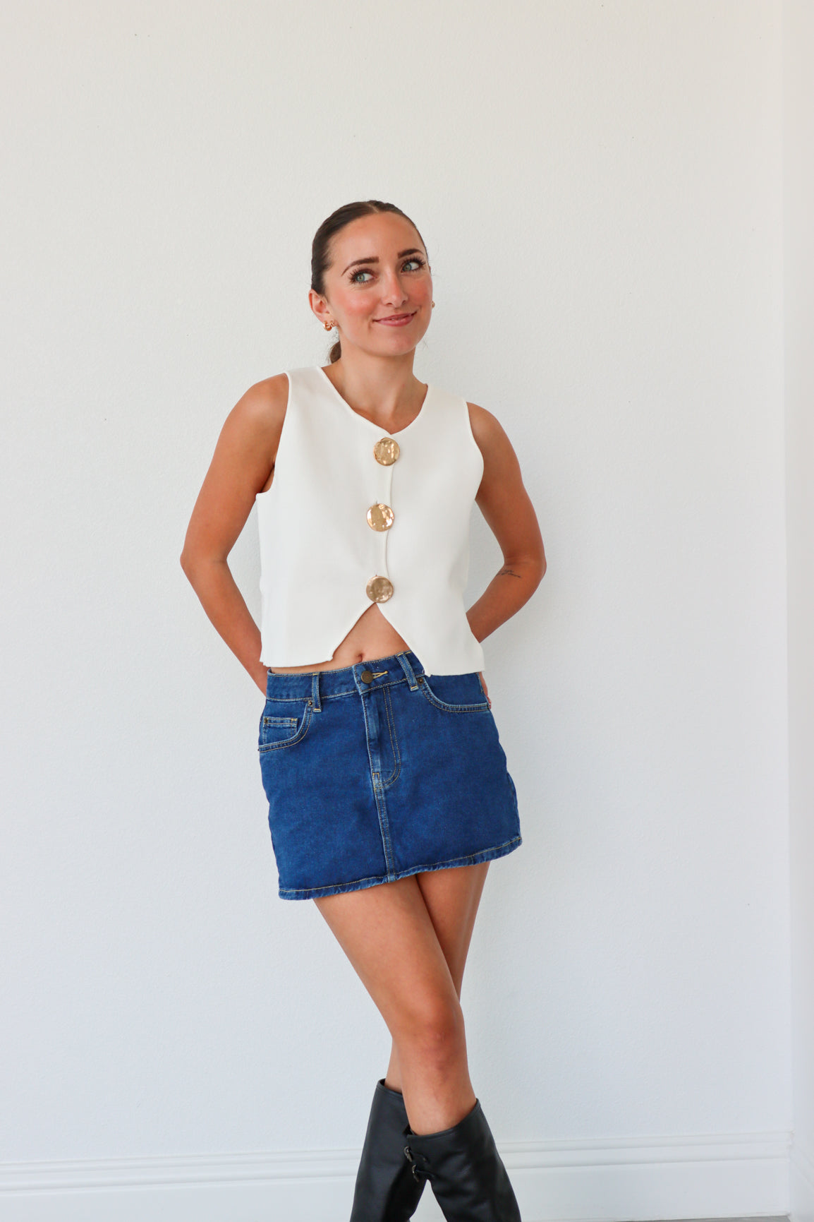 Woman wearing a white sleeveless top with gold buttons, blue denim skirt, and black knee-high boots against a white background.