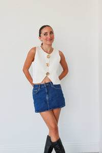 Woman wearing a white sleeveless top with gold buttons, blue denim skirt, and black knee-high boots against a white background.