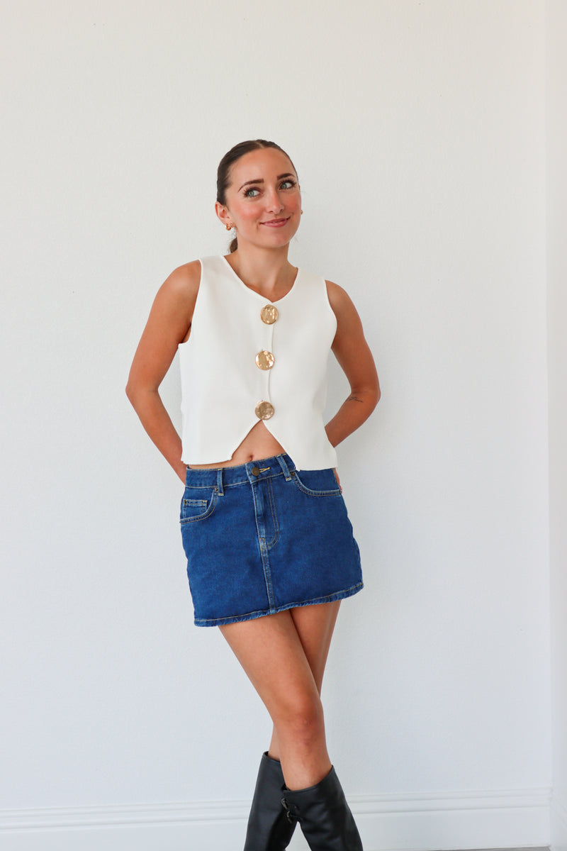 Woman wearing a white sleeveless top with gold buttons, blue denim skirt, and black knee-high boots against a white background.