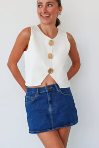 Woman wearing a white sleeveless top with gold buttons and a blue denim skirt against a white background