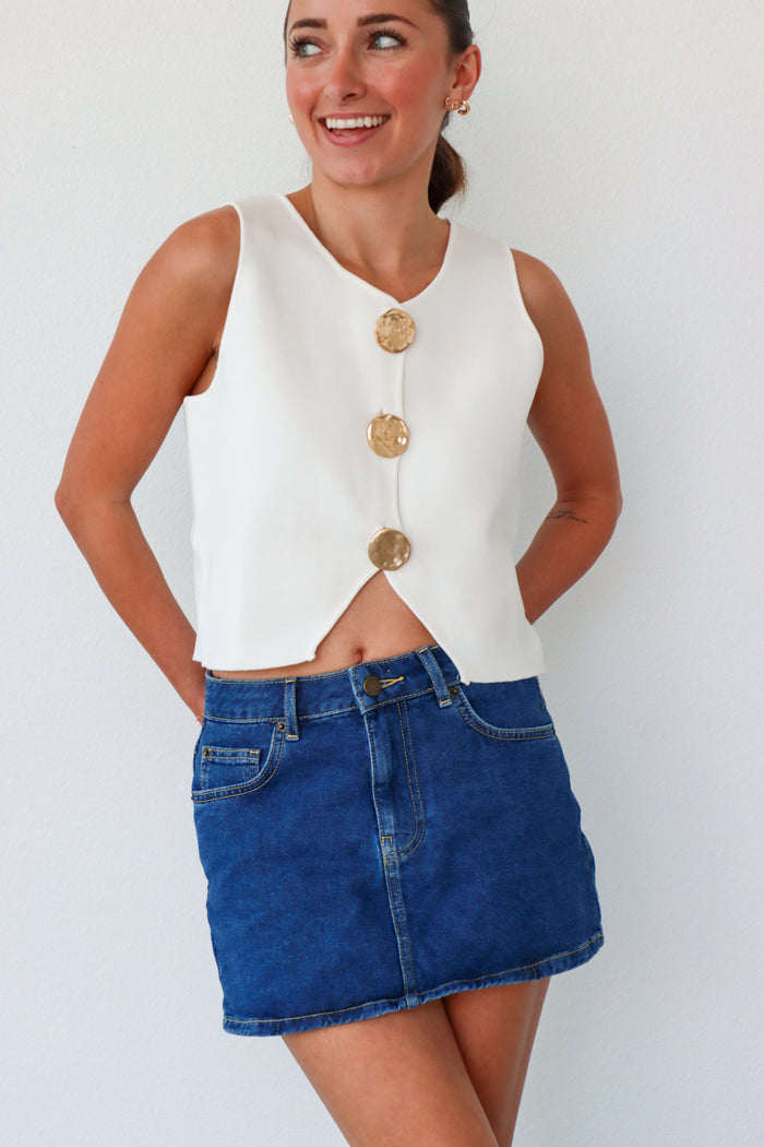Woman wearing a white sleeveless top with gold buttons and a blue denim skirt against a white background