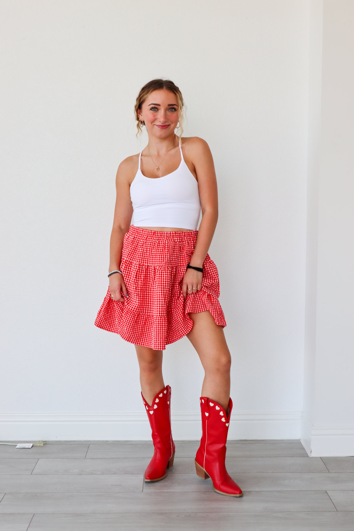 Woman wearing a white tank top, red checkered skirt, and red cowboy boots against a white wall.