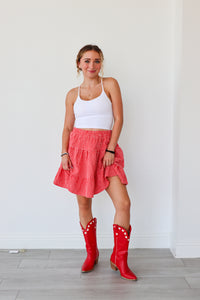 Woman wearing a white tank top, red checkered skirt, and red cowboy boots against a white wall.
