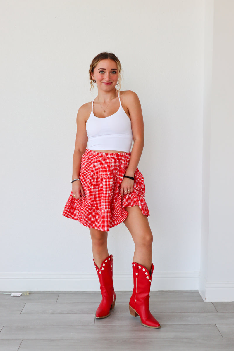 Woman wearing a white tank top, red checkered skirt, and red cowboy boots against a white wall.
